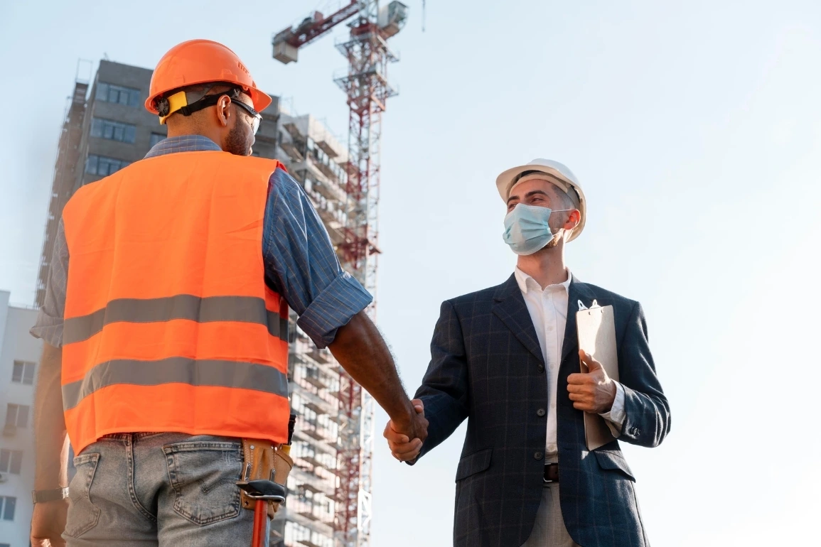 Two construction workers in safety gear examining blueprints on-site.